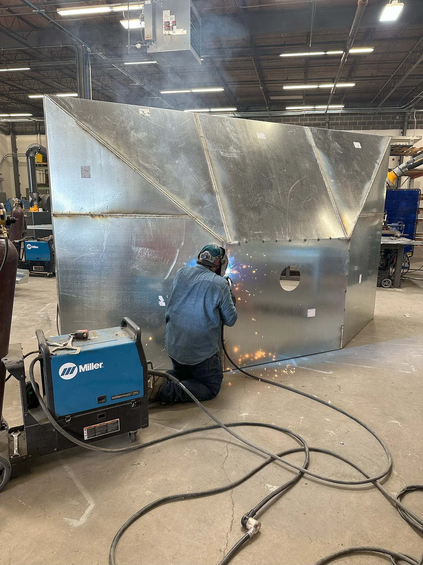 A welder using a Miller welding machine joins sections of large metal ductwork inside a fabrication shop, producing bright sparks during the process.