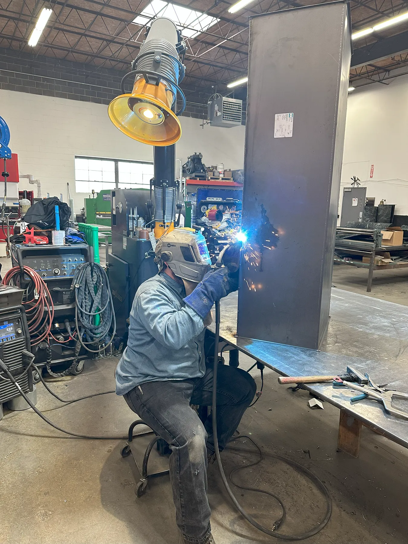 A professional welder wearing protective gear performs a precision weld on a large metal beam inside an industrial fabrication shop.